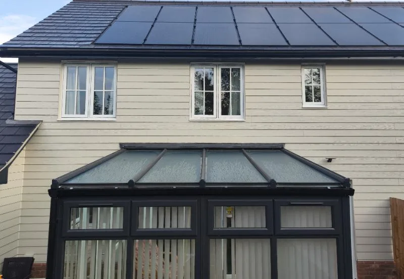 A conservatory with its original glazed roof, dark grey frames against a cream clad house, before an insulated panel roof was fitted.